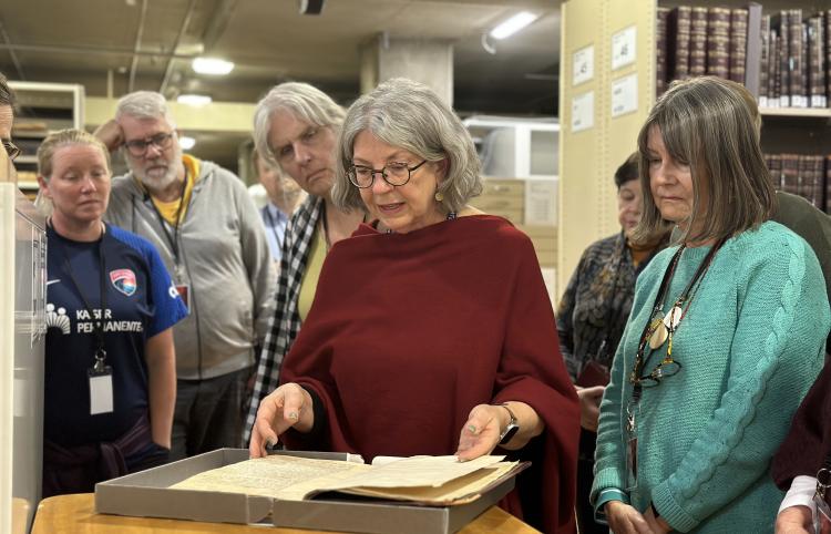A woman stands in front of a group of people, all gathered around and viewing a large old book.