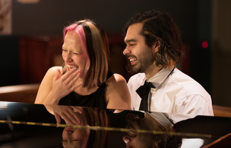 Two people sitting at a grand piano and laughing