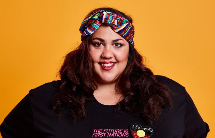 A smiling woman with long dark hair stands with her hands on her hips against a deep yellow background. She is wearing a black t-shirt that reads 'The Future is First Nations' in pink text, with an Aboriginal flag in the shape of a heart embroidered beside it. She wears a colourful headband.