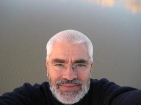 A man with white hair and glasses stands close to the camera with a pool of water behind him reflects the sky.