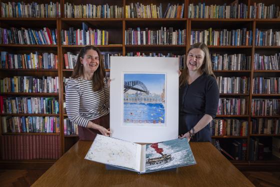 Two women stand in front of a bookshelf holding two colourful artworks depicting Sydney Harbour scenes.