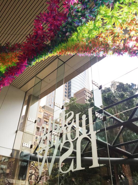 A colourful plastic sculpture of flowers, hangs from the ceiling - the sign on the window below reads State Library of NSW