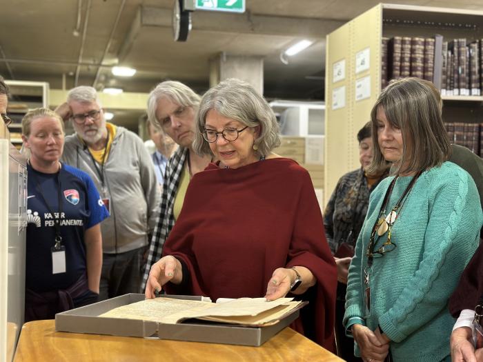 A woman stands in front of a group of people, all gathered around and viewing a large old book.