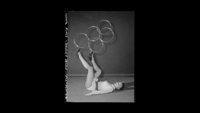 A black and white photo of a rhythmic gymnast in a leotard and high heels lies on her back, balancing 5 silver rings in the Olympics logo formation on her feet. White handwriting around the left edge of the photo says 'OLYMPIC RINGS - SHIRLEY REGAN - "PIX" OCT '56'