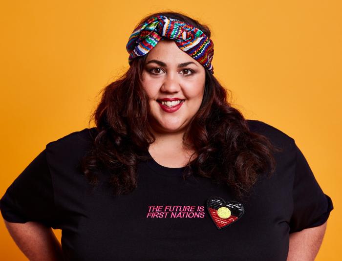 A smiling woman with long dark hair stands with her hands on her hips against a deep yellow background. She is wearing a black t-shirt that reads 'The Future is First Nations' in pink text, with an Aboriginal flag in the shape of a heart embroidered beside it. She wears a colourful headband.